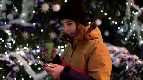 Junge-Frau-mit-Tasse-heißen-Tee-mit-ihrem-Smartphone-Stand-in-der-Nähe-der-Weihnachtsbaum.