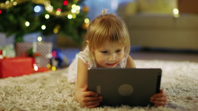 Niedliche-kleine-Mädchen-mit-Tablet-Computer-in-ihren-Händen-liegt-auf-dem-Teppich-unter-dem-Weihnachtsbaum.
