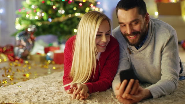 Glückliches-Paar-liegt-auf-dem-Teppich-unter-Weihnachtsbaum,-schauen-Sie-sich-etwas-Interessantes-auf-dem-Smartphone.