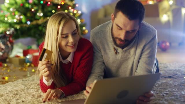 Brautpaar-auf-dem-Teppich-unter-dem-Weihnachtsbaum-liegen,-Frau-hält-Kreditkarte-und-Mann-kauft-ihr-Geschenke-auf-dem-Laptop.