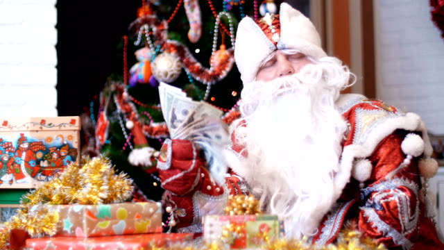 Santa-Claus,-Weihnachtsmann,-Väterchen-Frost-fächelt-sich-mit-einem-Fan-von-Dollar,-er-sitzt-an-einem-Tisch-unter-die-Weihnachtsgeschenke,-im-Hintergrund-ist-ein-Weihnachtsbaum