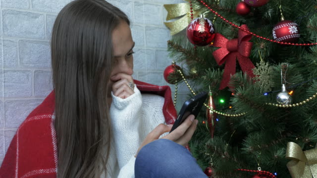 Frau-sitzt-auf-einer-Etage-mit-roten-Plaid-und-mit-Smartphone-neben-Weihnachtsbaum