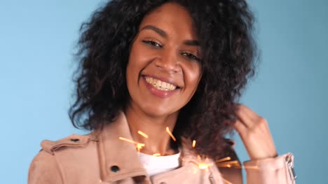 Sparkling-Bengal-fire-in-african-american-woman's-hand-on-blue-background.-Christmas-Holiday-Concept.-Young-pretty-girl-with-afro-hairstyle-celebrating,-smiling,-enjoying-time.-4k