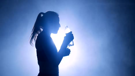Thirsty-female-boxer-taking-a-break-drinking-from-the-water-bottle-after-training-in-the-dark-gym-with-smoke.-Silhouettes-of-a-woman-boxer-in-dark-gym