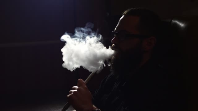 junge-bärtiger-Mann-in-Gläsern-raucht-eine-Wasserpfeife-und-Ausblasen-Rauch-Closeup-auf-schwarzem-Hintergrund-in-Zeitlupe-in-4k