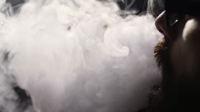 young-bearded-man-in-glasses-smokes-a-hookah-and-blow-out-smoke-closeup-on-black-background-in-slow-motion-in-4k
