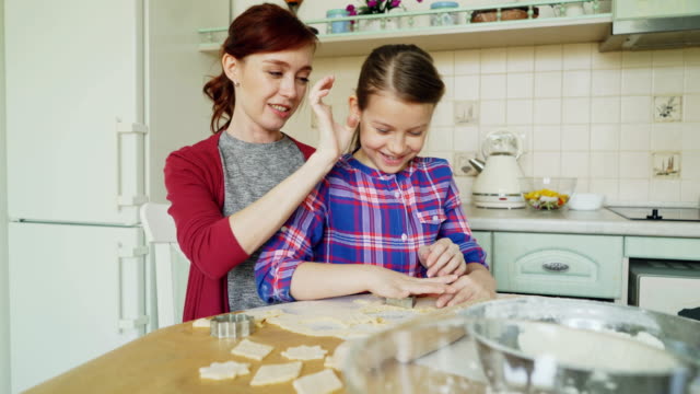 Fröhliche-Mutter-und-kleine-niedliche-Tochter-sprechen-und-Cookies-zusammen-mit-Bäckerei-Formen-Zuschneiden-Teig-während-der-Sitzung-in-der-modernen-Küche-zu-Hause-zu-machen.-Familie,-Essen-und-Leute-Konzept