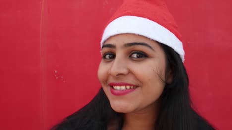 Young-Indian-woman-with-Santa's-hat-welcome-and-making-hand-gestures,-excited-and-super-happy,-with-a-matching-red-background,-handheld-gimbal-stabilized