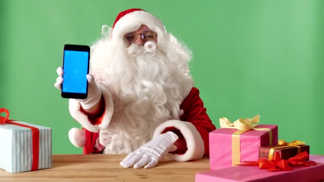 Sonriendo-Santa-Claus-mostrando-en-cámara-smartphone,-regalos-sobre-la-mesa,-chromakey-en-el-fondo