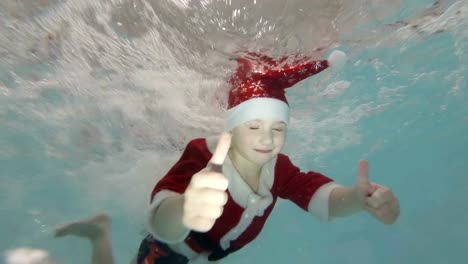 Niño-feliz-en-un-traje-rojo-de-Santa-Claus-se-presenta-bajo-el-agua-en-la-piscina,-jugando-en-los-chorros-de-agua,-sonriendo-y-mirando-a-la-cámara.
