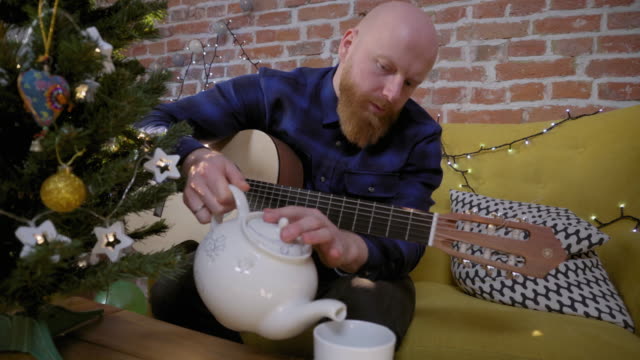 Man-with-guitar-sitting-next-to-Christmas-tree-and-pouring-tea
