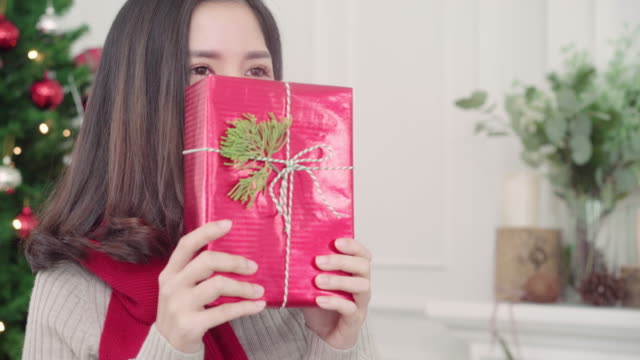 Fröhlich-glücklich-junge-Asiatin-hält-lächelnd-zu-Kamera-in-ihrem-Wohnzimmer-zu-Hause-in-Weihnachtsfest-Weihnachtsgeschenke.-Lifestyle-Frau-feiern-Weihnachten-und-Neujahr-Konzept.