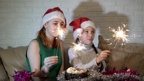 Happy-children-in-Santa's-hats-with-lighted-sparklers-play-sitting-on-the-couch-near-the-cake-and-smile.