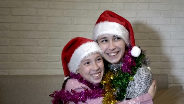 Two-cute-young-girls-in-Santa-Claus-hats-and-Christmas-tinsel-on-their-shoulders-hugging-sitting-on-the-couch,-laughing-and-looking-at-the-camera.-Portrait.-Close-up.-4K.-25-fps.