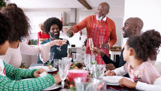 Senior-schwarzer-Mann-trägt-eine-Schürze-gießt-Champagner-für-seine-Familie-am-Esstisch-zu-feiern,-während-Weihnachtsessen-vorbereitet,-selektiven-Fokus