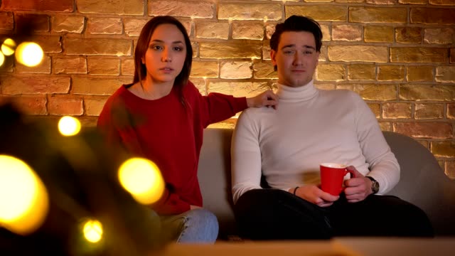 Umarmen-kaukasischen-paar-sitzt-auf-Sofa-und-aufmerksam-beobachten-Film-mit-Tasse-Getränk-in-weihnachtlicher-Atmosphäre.