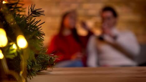 Árbol-de-Navidad-con-luces-y-borrosa-pareja-de-jóvenes-sentados-en-sofá-y-hablando-en-el-fondo.