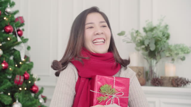 Fröhlich-glücklich-junge-Asiatin-hält-lächelnd-zu-Kamera-in-ihrem-Wohnzimmer-zu-Hause-in-Weihnachtsfest-Weihnachtsgeschenke.-Lifestyle-Frau-feiern-Weihnachten-und-Neujahr-Konzept.