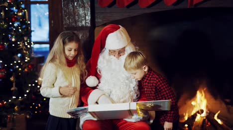 Santa-Claus-is-sitting-in-the-middle-of-a-room-with-an-album-in-his-hands,-surrounded-by-children-on-the-background-of-a-fireplace,-and-telling-a-New-Year-story.
