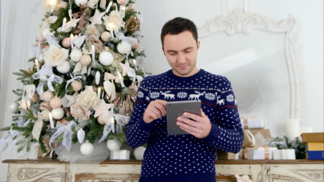 Junger-Mann-in-Weihnachtsstrickbekleidung-überprüft-sein-Tablet