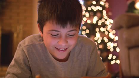 Niño-tendido-en-el-suelo-usando-su-tableta,-sonríe-divertido-a-la-cámara,-árbol-de-Navidad-con-luces-detrás-de-él