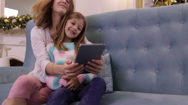 Madre-con-hija-en-el-sofá-usando-la-tableta-computadora-feliz-sonrisa-joven-familia-cerca-decorada-año-nuevo-árbol-de-Navidad