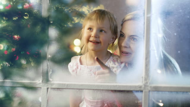 On-Christmas-Eve-Mother-and-Daughter-Looking-Through-Snowy-Window.-Garland-Shines-Bright-on-a-Window.