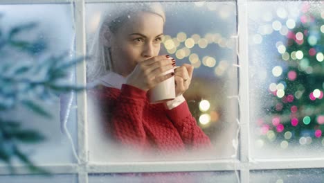 En-Nochebuena-hermosa-mujer-está-mirando-por-la-ventana-Nevada.-Ella-está-sosteniendo-una-taza-con-una-bebida-caliente-y-se-siente-acogedor.