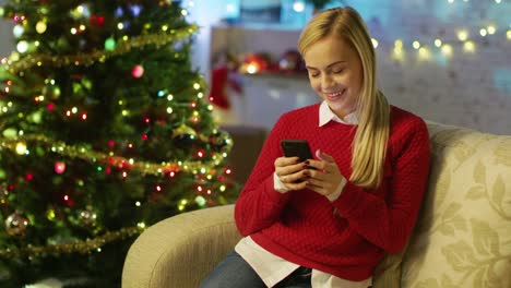 Schöne-Blonde-Frau-sitzt-auf-einer-Couch-und-Smartphone.-Weihnachtsbaum-und-Zimmer-mit-Lichtern-geschmückt-werden-im-Hintergrund.
