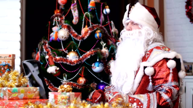 Santa-Claus,-Weihnachtsmann,-Väterchen-Frost-arbeitet-an-einem-Laptop-an-einem-Tisch,-unter-Weihnachten-präsentiert,-im-Hintergrund-ist-Weihnachtsbaum