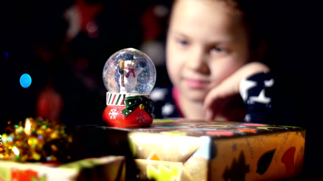 In-the-twilight-of-the-night,-among-the-gifts-in-bright-colorful-paper-packages,-a-pretty-little-blonde-girl-with-a-pink-bow-in-her-hair-and-a-beautiful-dress,-looks-carefully-and-admires-snow-globe.-toy-Christmas-ball-with-sparkles-and-a-snowman-inside