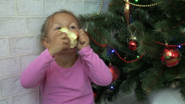 Cute-niña-sentada-en-el-suelo-y-comer-manzana-al-lado-de-un-árbol-de-Navidad.