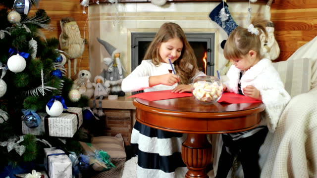children-write-Letter-for-Santa-Claus,-sisters-eating-sweets,-girls-sit-near-the-fireplace,-children-near-Christmas-tree,-letter-to-Santa