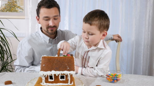 Little-son-with-father-decorates-a-gingerbread-house-with-a-colorful-candies