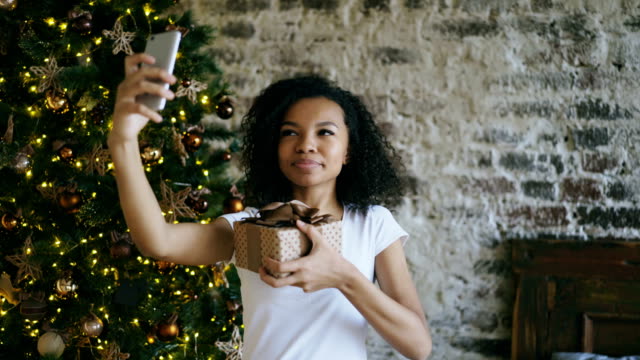 Chica-divertida-carrera-mixta-selfie-fotografiando-en-la-cámara-del-smartphone-en-el-país-cerca-de-árbol-de-Navidad