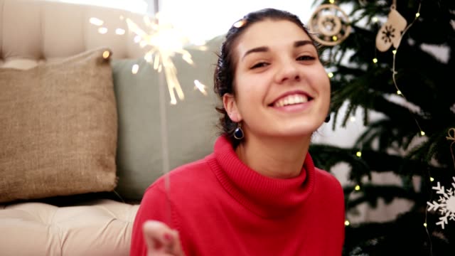 Feliz-atractiva-mujer-joven-sentada-en-el-piso-por-el-árbol-de-Navidad-celebrando-Navidad-con-luces-de-Bengala-y-mirando-la-cámara