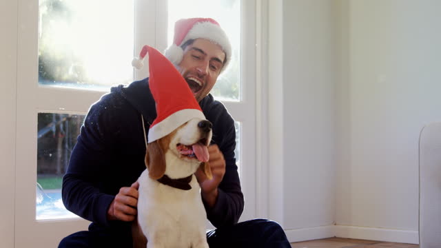 Smiling-young-man-putting-santa-cap-on-his-pet-dog-4K-4k