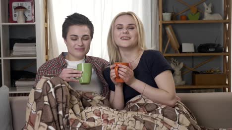 Two-young-lesbian-girls-are-sitting-on-the-couch,-covered-with-a-warm-blanket,-holding-cups-in-their-hands,-drinking-dark-tea,-coffee,-cuddling-60-fps