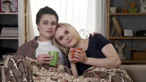 Two-young-lesbian-girls-are-sitting-on-the-couch,-covered-with-a-warm-blanket,-holding-cups-in-their-hands,-cuddling,-looking-at-the-camera-60-fps