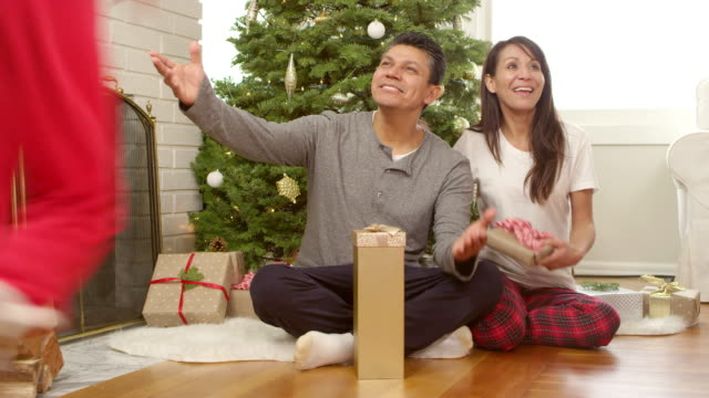 A-man-and-woman-sit-in-front-of-a-christmas-tree-with-presents-and-are-then-are-joined-by-their-two-daughters