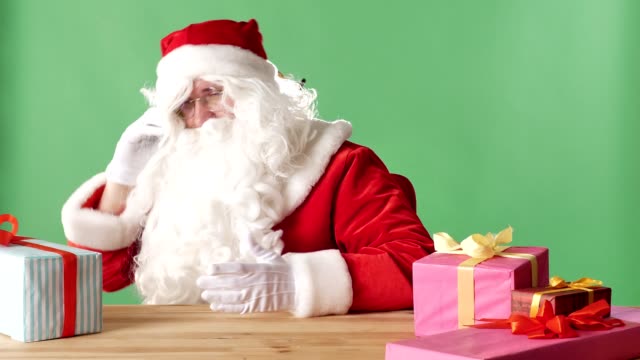 Satisfied-Santa-Claus-speaks-on-the-phone,-laughs,-sits-at-table-with-gifts,-green-chromakey-in-the-background