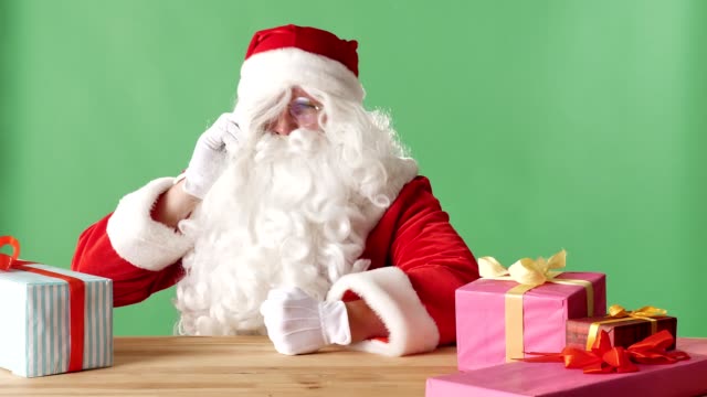 Böser-Weihnachtsmann-am-Telefon-spricht,-schlägt-seine-Faust-auf-den-Tisch,-sitzt-am-Tisch-mit-Geschenken,-grüne-Chromakey-im-Hintergrund