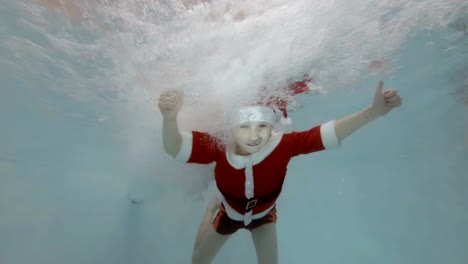 Un-poco-chico-nada-bajo-el-agua-en-los-chorros-de-agua-en-la-piscina-con-un-traje-de-Santa-Claus-con-sus-brazos-extendidos,-sonríe,-mira-a-la-cámara-y-demuestra-sus-dedos-para-arriba.-Cámara-lenta.