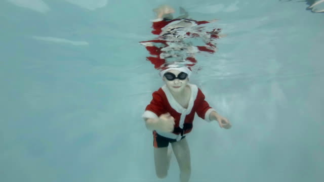 A-happy-little-boy-in-a-suit-and-a-red-Santa-Claus-hat-is-submerged-in-the-pool.-The-boy-is-swimming,-smiling-and-looking-into-the-camera.