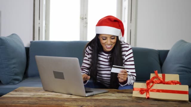 Frau-mit-roten-Pullover-und-Weihnachtsmann-Mütze-mit-Kreditkarte-auswählen-und-kaufen-Weihnachtsgeschenke-mit-Laptop-zu-Hause-begeistert-mit-Verkauf-über-das-Internet-und-Kreditkarte-Einrichtungen-in-Linie-einkaufen