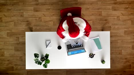 Santa-Claus-im-Büro-am-Laptop-arbeiten,-schreiben,-trinken-Kaffee,-Luftaufnahme,-Top-down-Schuss