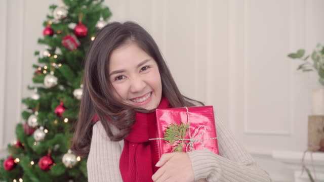 Cheerful-happy-young-Asian-woman-holding-christmas-gifts-smiling-to-camera-in-her-living-room-at-home-in-Christmas-Festival.-Lifestyle-woman-celebrate-Christmas-and-New-year-concept.