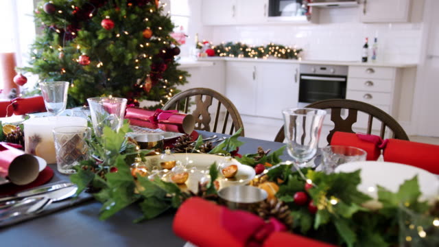 Eine-dekorierte-Weihnachtstisch-mit-Christmas-Cracker-auf-Platten-in-einem-Speisesaal-angeordnet,-ein-Weihnachtsbaum-mit-Küche-im-Hintergrund