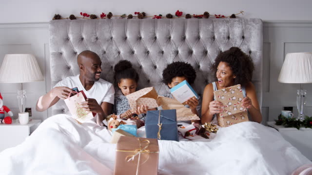 Jóvenes-mixtos-familia-carrera-sentado-en-la-cama-juntos-desenvolver-regalos-en-la-mañana-de-Navidad,-vista-frontal