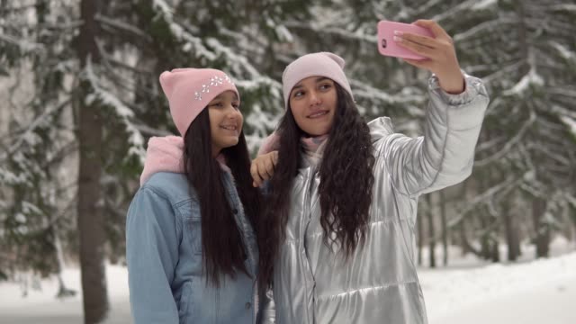 Two-young-girlfriends-in-winter-clothes-taking-selfie-against-the-background-of-the-winter-forest-using-a-smartphone-and-smiling.